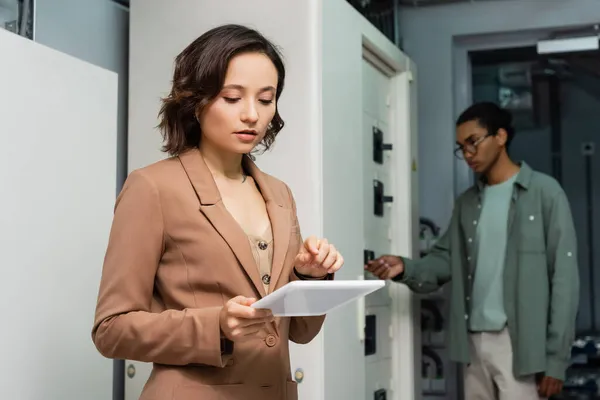Young engineer pointing at digital tablet while blurred african american colleague checking switchboard — Stock Photo
