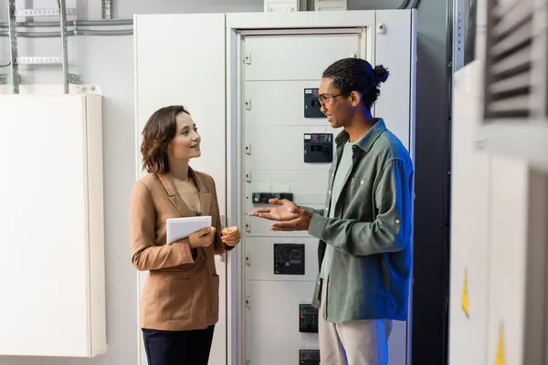 Afrikanischer amerikanischer Ingenieur zeigt mit den Händen, während er mit einem Kollegen mit digitalem Tablet in der Nähe der Schalttafel spricht — Stockfoto