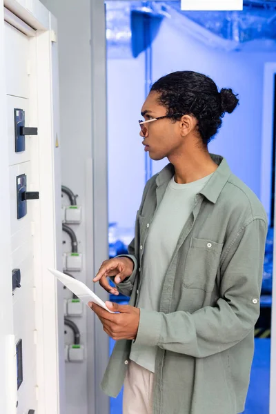 Ingeniero afroamericano apuntando a la tableta digital mientras comprueba la centralita en el centro de datos - foto de stock