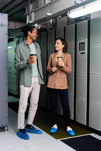 Ingenieros interracial sonrientes sosteniendo café para llevar durante la conversación en el centro de datos - foto de stock