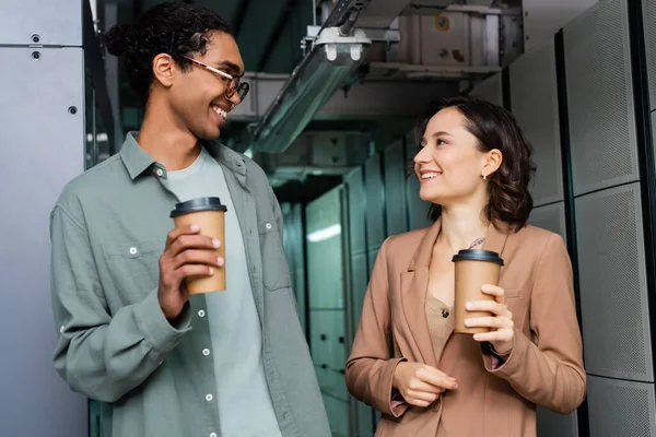Happy interracial programmers with paper cups talking in data center — Stock Photo