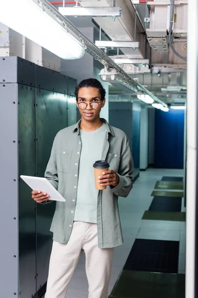 Positive african american administrator with digital tablet and coffee to go looking at camera in data center — Stock Photo