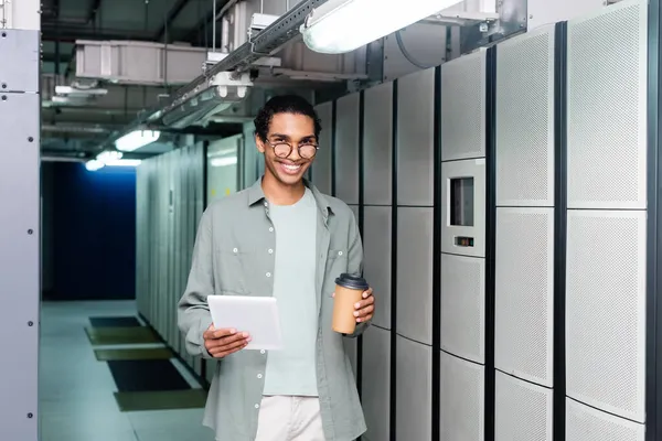 Happy african american engineer with digital tablet and coffee to go looking at camera in data center — Stock Photo