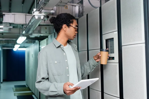 Tecnico afroamericano con caffè da andare e tablet digitale toccando il pannello di controllo sul server — Foto stock