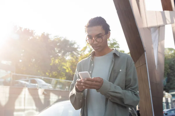 Trendy african american man in eyeglasses messaging on mobile phone outdoors — Stock Photo