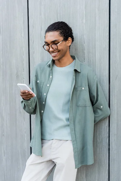 Sonriente africano americano hombre charlando en el teléfono móvil mientras de pie wth mano detrás de nuevo cerca de gris pared - foto de stock