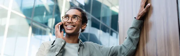 Joyeux homme afro-américain regardant loin tout en parlant sur un téléphone mobile à l'extérieur, bannière — Photo de stock