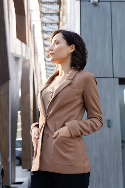 Jeune femme regardant loin tout en se tenant avec les mains dans des poches de blazer beige à l'extérieur — Photo de stock