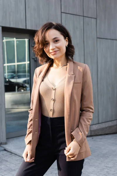 Morena mujer en chaqueta beige sonriendo a la cámara mientras está de pie al aire libre - foto de stock