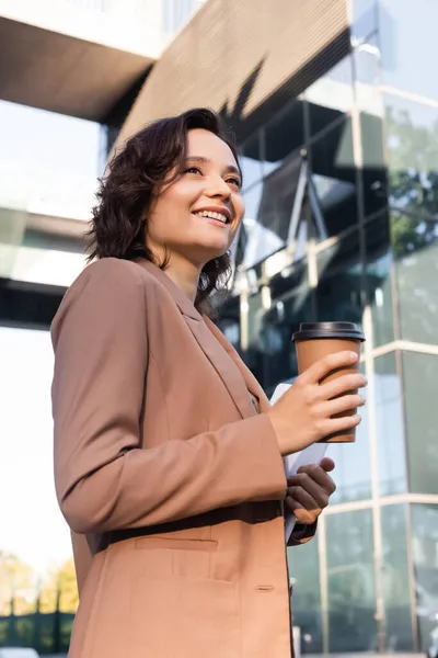 Baixo ângulo vista de mulher elegante com café para ir sorrindo enquanto olha para o exterior — Fotografia de Stock