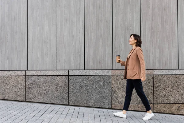 Вид сбоку стильной женщины, идущей с кофе вдоль серой стены — стоковое фото