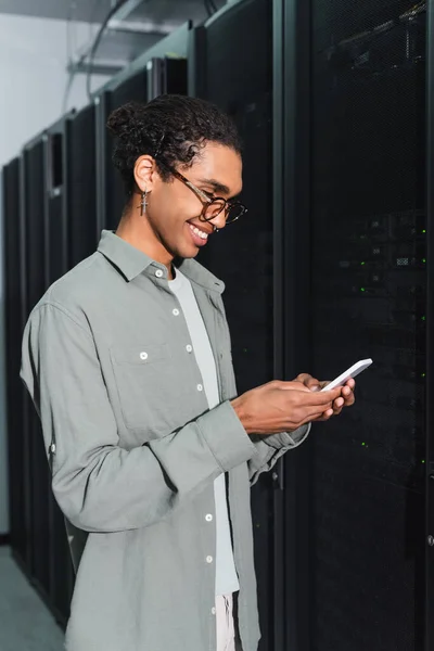 Mensagens de programador americano africano alegre no telefone celular perto do servidor no data center — Fotografia de Stock