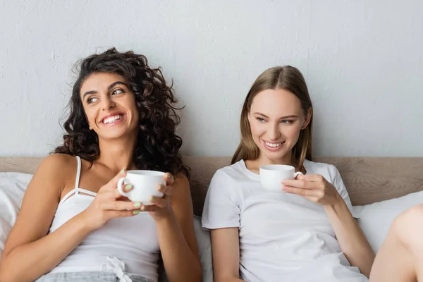 Счастливая лесбийская пара держит чашки кофе в спальне — стоковое фото