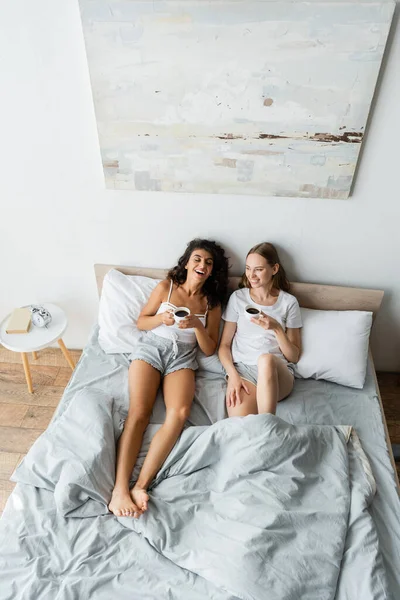 Vue grand angle de couple lesbienne heureux tenant des tasses de café au lit — Photo de stock