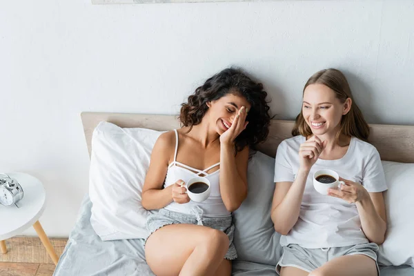 Vue grand angle de joyeux couple lesbien tenant tasses de café au lit — Photo de stock