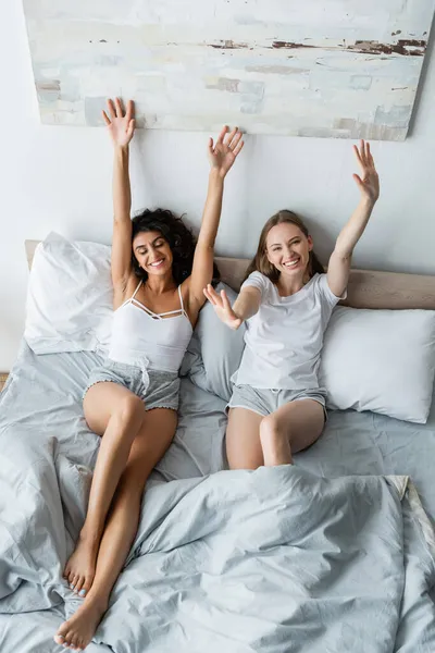 Vue grand angle de joyeux couple lesbien couché avec les mains levées dans le lit — Photo de stock