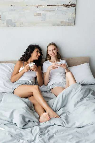Pleased young lesbian couple holding cups of coffee in bed — Stock Photo