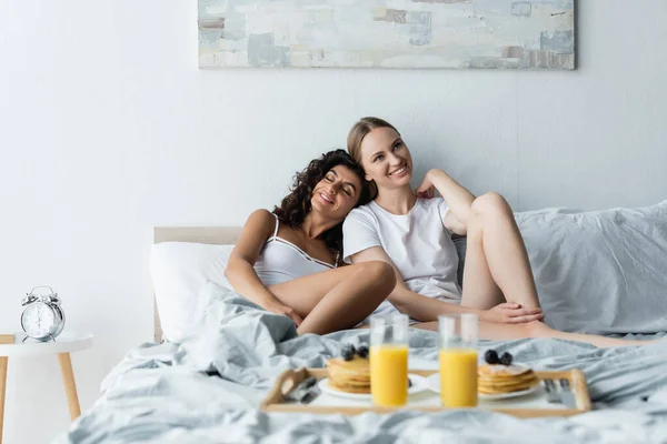 Feliz joven mujer apoyada en novia cerca de bandeja con desayuno en la cama - foto de stock