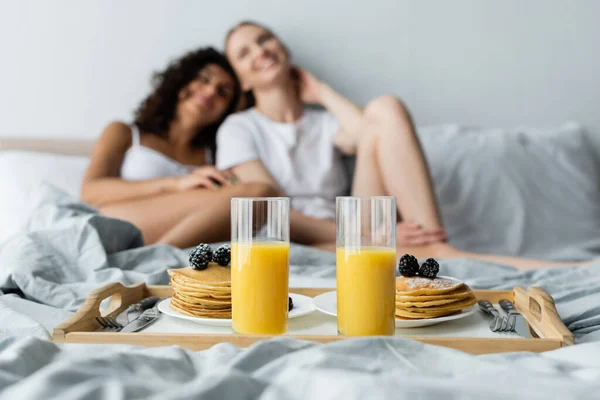 Óculos com suco de laranja e panquecas na bandeja perto de casal de lésbicas borradas — Fotografia de Stock