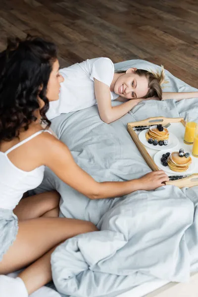 Femme heureuse couché et regardant petite amie frisée près du plateau avec petit déjeuner sur le lit — Photo de stock