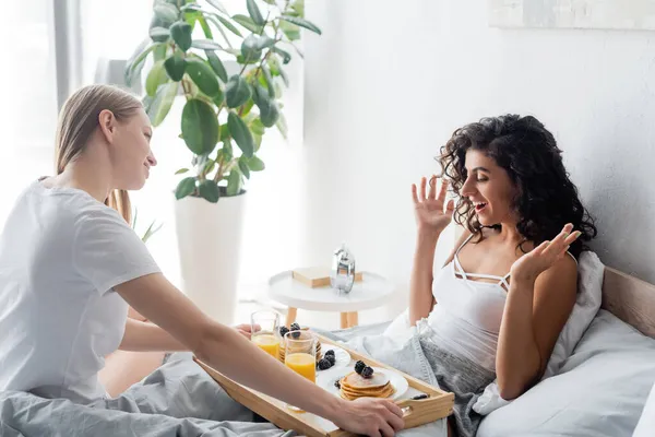 Young woman bringing tray with breakfast to excited girlfriend in bed — Stock Photo