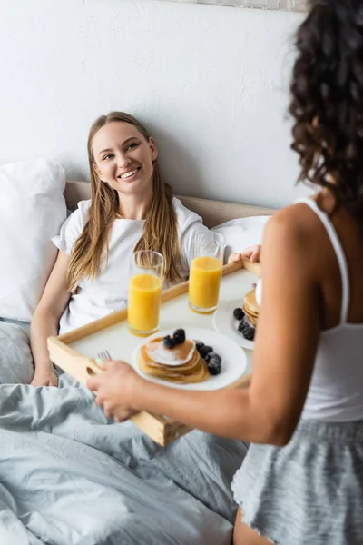 Verschwommene Frau hält Tablett mit Frühstück neben glücklicher Freundin im Bett — Stockfoto