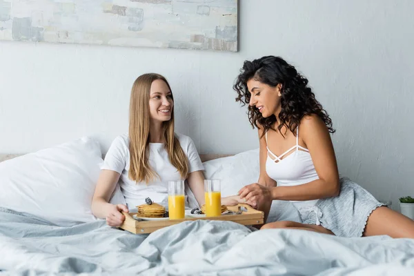 Счастливая лесбийская пара улыбается возле подноса для завтрака на кровати — стоковое фото