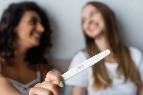 Mujer borrosa y alegre sosteniendo prueba de embarazo cerca de novia feliz - foto de stock