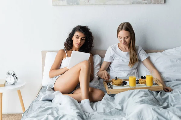 Femme bouclée utilisant tablette numérique près de petite amie ayant le petit déjeuner au lit — Photo de stock