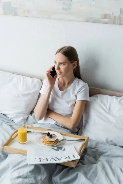 Mujer joven hablando en teléfono inteligente cerca de la bandeja con desayuno en el dormitorio - foto de stock