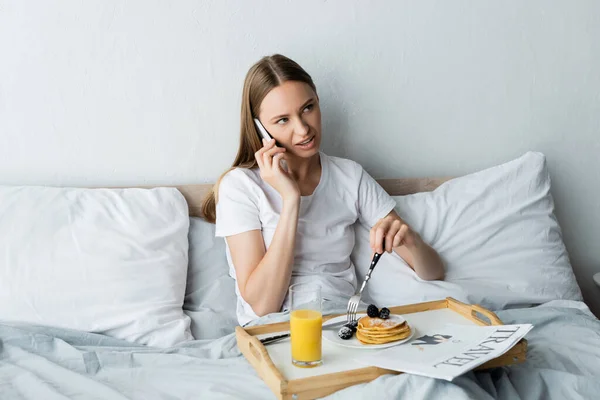 Jovem mulher falando no smartphone perto da bandeja com café da manhã no quarto — Fotografia de Stock