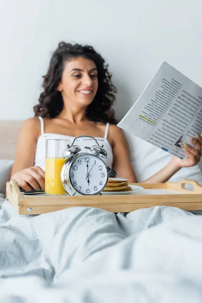 Happy and blurred woman reading travel newspaper near tray with alarm clock and breakfast — Stock Photo