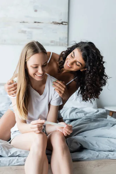 Cheerful woman holding pregnancy test while hugging joyful girlfriend in bedroom — Stock Photo