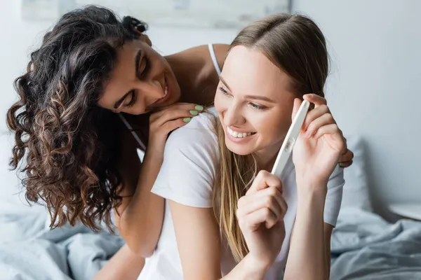 Happy woman holding pregnancy test while hugging joyful girlfriend in bedroom — Stock Photo