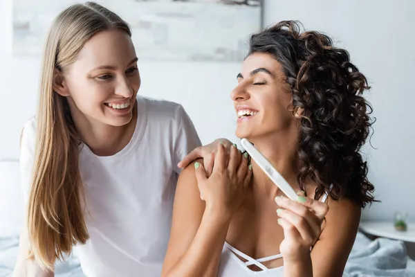 Mujer feliz y rizada sosteniendo prueba de embarazo cerca de novia en el dormitorio - foto de stock