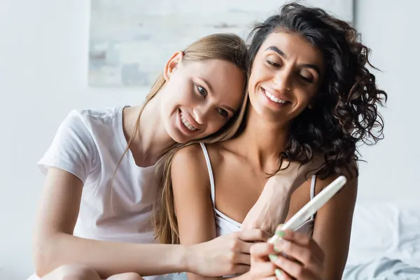 Mujer alegre celebración de prueba de embarazo mientras se abraza con novia feliz en el dormitorio - foto de stock