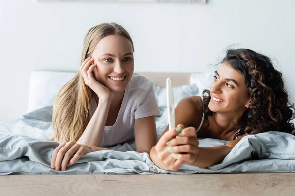 Cheerful woman holding pregnancy test near happy girlfriend in bedroom — Stock Photo