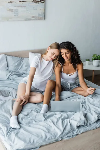 Feliz pareja de lesbianas mirando a la computadora portátil, mientras que las compras en línea y sentado en la cama - foto de stock