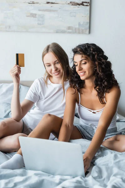 Улыбающаяся лесбийская пара смотрит на ноутбук во время покупок в спальне — стоковое фото