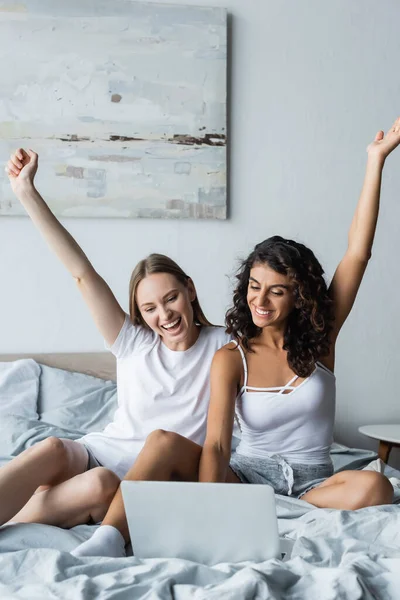 Excited lesbian couple with raised hands looking at laptop on bed — Stock Photo