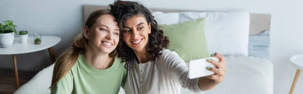 Curly woman taking selfie with happy girlfriend in bedroom, banner — Stock Photo