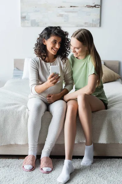 Schwangere hält Smartphone in der Hand und sitzt mit glücklicher Freundin im Bett — Stockfoto