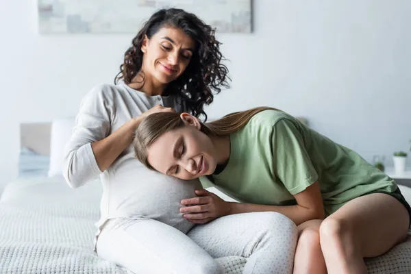 Happy woman listening sound of belly near pregnant girlfriend in bedroom — Stock Photo