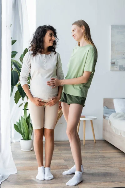 Full length of happy woman touching belly of pregnant girlfriend in bedroom — Stock Photo