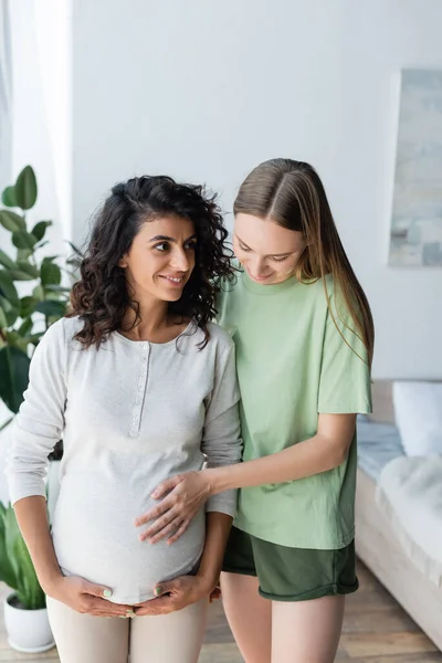 Glückliche Frau berührt Bauch einer schwangeren Freundin, während sie im Schlafzimmer steht — Stockfoto