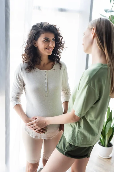 Young woman touching belly of happy pregnant girlfriend in bedroom — Stock Photo