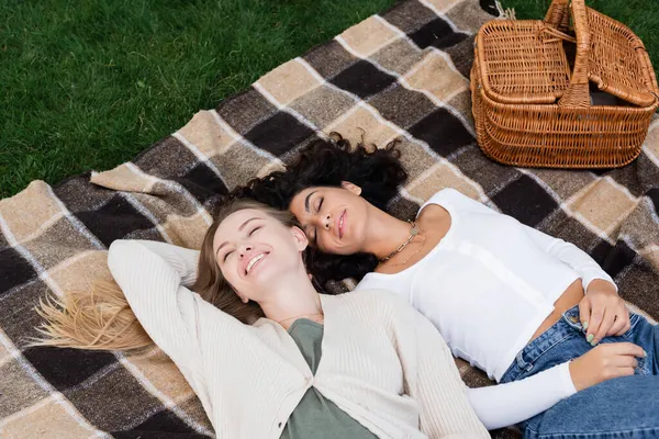 Vue grand angle du couple lesbienne heureux couché sur une couverture à carreaux pendant le pique-nique — Photo de stock