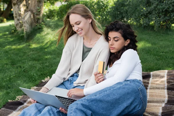 Curly woman holding credit card while doing online shopping with happy girlfriend during picnic — Stock Photo