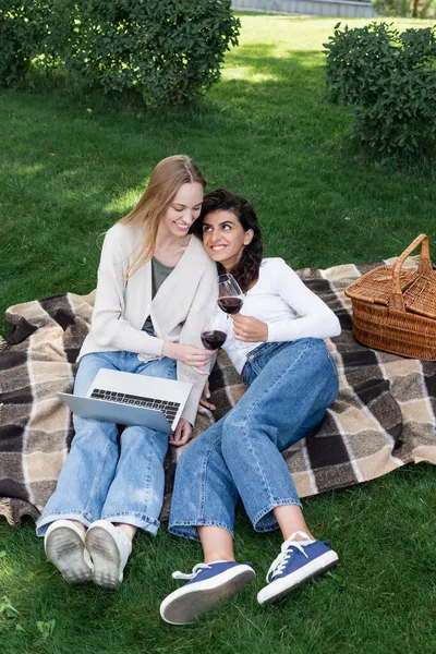 Femmes heureuses tenant des verres de vin rouge près d'un ordinateur portable pendant le pique-nique — Photo de stock