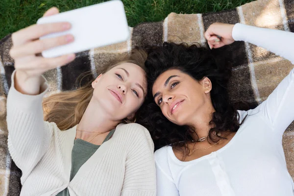 Blick aus der Vogelperspektive auf glückliche Frauen, die ein Selfie machen, während sie auf einer Decke im Park liegen — Stockfoto